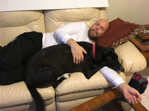 Paul is lying on a couch with Blackie, his dog, who has her head on Paul's arm.